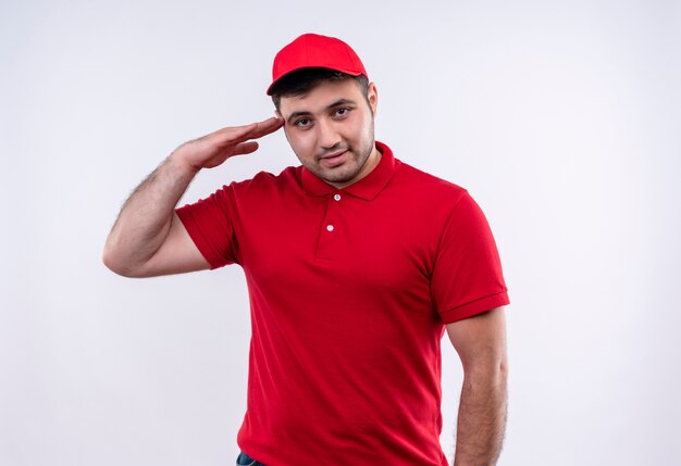 Young delivery man in red uniform and cap smiling confident saluting standing over white wall