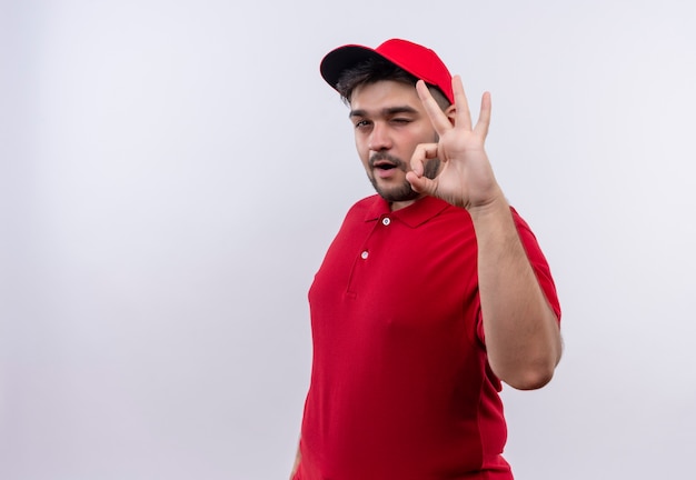 Young delivery man in red uniform and cap smiling confident winking showing ok sign 