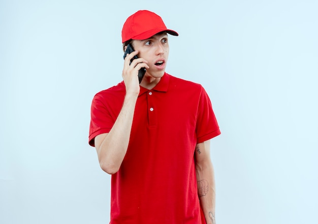 빨간 제복을 입은 젊은 배달 남자와 휴대 전화에 얘기하는 모자는 흰 벽에 놀라고 놀란 찾고