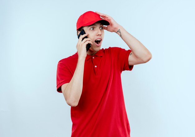 Young delivery man in red uniform and cap talking on mobile phone looking surprised and amazed standing over white wall