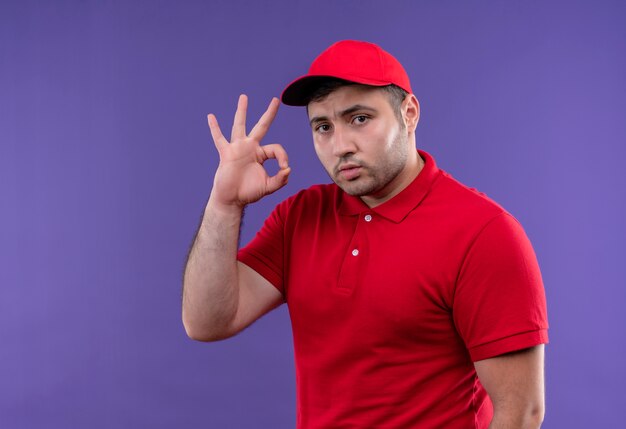 Young delivery man in red uniform and cap with confident expression doing ok sing standing over purple