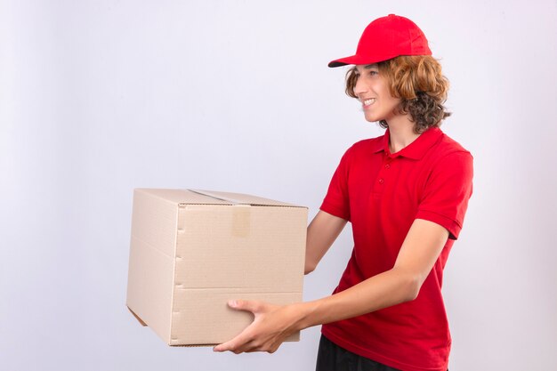 Young delivery man in red uniform giving cardboard box to a customer smiling friendly over isolated white background