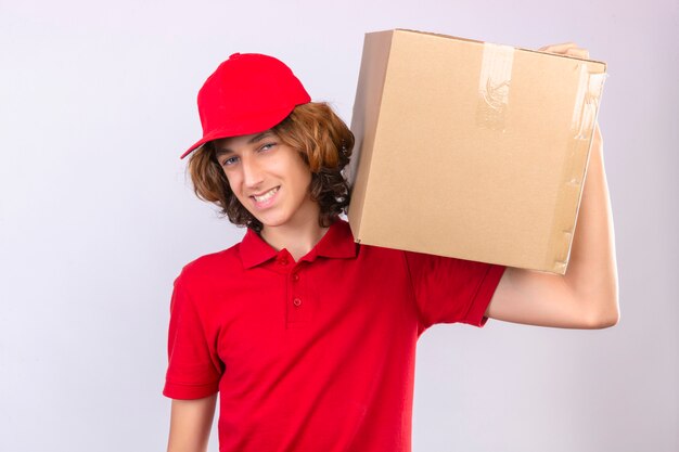 Молодой курьер в красной форме держит картонную коробку на плече, глядя в камеру, дружелюбно улыбаясь на изолированном белом фоне