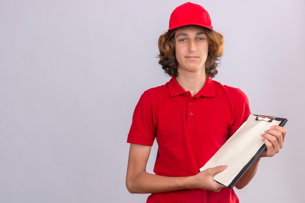 Foto gratuita giovane uomo di consegna in uniforme rossa che tiene appunti che guarda l'obbiettivo con la faccia seria sopra fondo bianco isolato