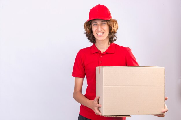 Young delivery man in red uniform holding large cardboard box looking at camera smiling cheerfully standing over isolated white background