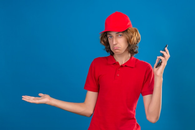 빨간색 제복을 입은 젊은 배달 남자가 손에 휴대 전화를 우둔하고 고립 된 파란색 배경 위에 서있는 두 팔을 벌려 혼란스러워합니다.