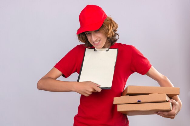 Foto gratuita giovane uomo di consegna in uniforme rossa che tiene scatole per pizza e appunti per chiedere la firma su bianco su sfondo bianco isolato