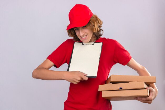 Молодой курьер в красной форме держит коробки для пиццы и буфер обмена с просьбой поставить подпись на бланке на изолированном белом фоне