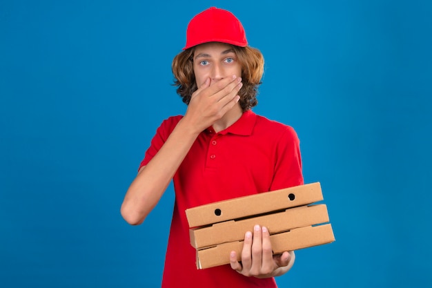 Young delivery man in red uniform holding pizza boxes looking surprised covering mouth with hand over isolated blue background