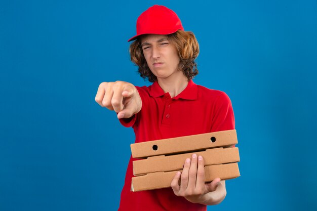 Young delivery man in red uniform holding pizza boxes pointing displeased and frustrated to the camera angry and furious with you over isolated blue background