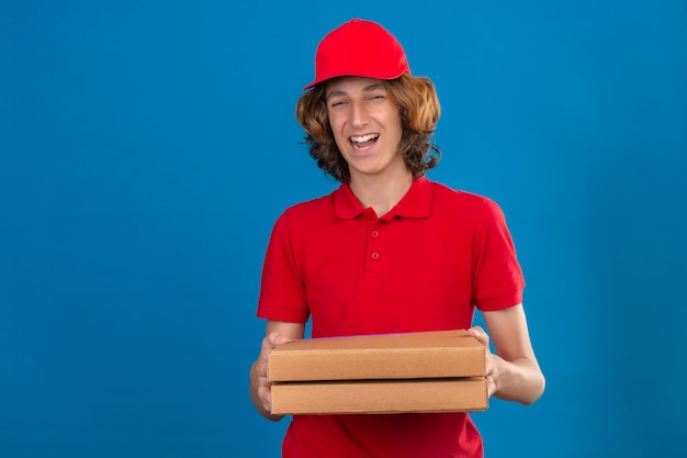 Young delivery man in red uniform holding pizza boxes smiling cheerfully with happy face over isolated blue background