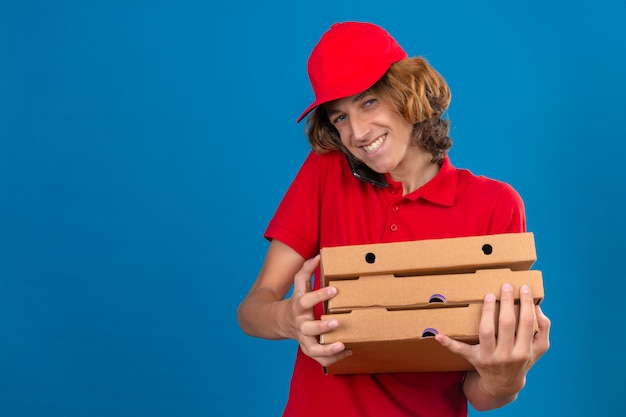 Young delivery man in red uniform holding pizza boxes while talking on mobile phone smiling with happy face over isolated blue background