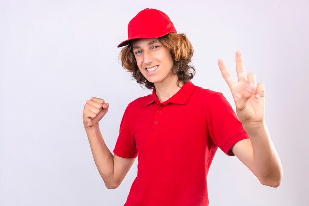 Young delivery man in red uniform looking at camera with happy face raising fist showing victory sign with fingers over isolated white background