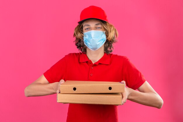 Young delivery man in red uniform wearing medical mask holding pizza boxes looking at camera smiling friendly with happy face over isolated pink background