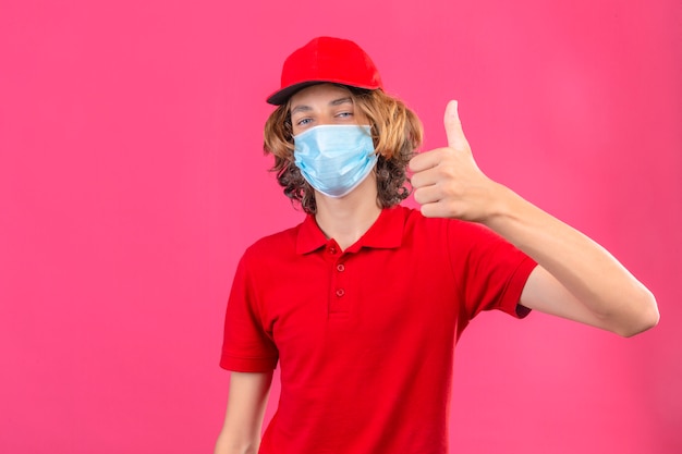 격리 된 분홍색 배경 위에 유쾌 하 게 엄지를 보여주는 미소를 카메라를보고 의료 마스크를 쓰고 빨간 제복을 입은 젊은 배달 남자