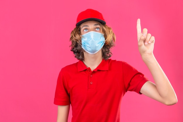 Young delivery man in red uniform wearing medical mask looking up pointing with finger standing over isolated pink background