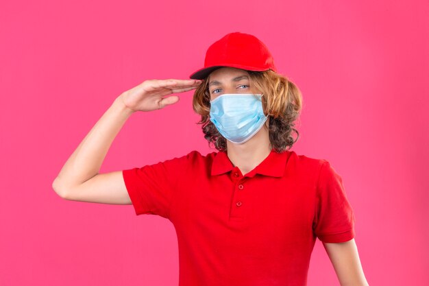 Young delivery man in red uniform wearing medical mask saluting looking confident standing over isolated pink background