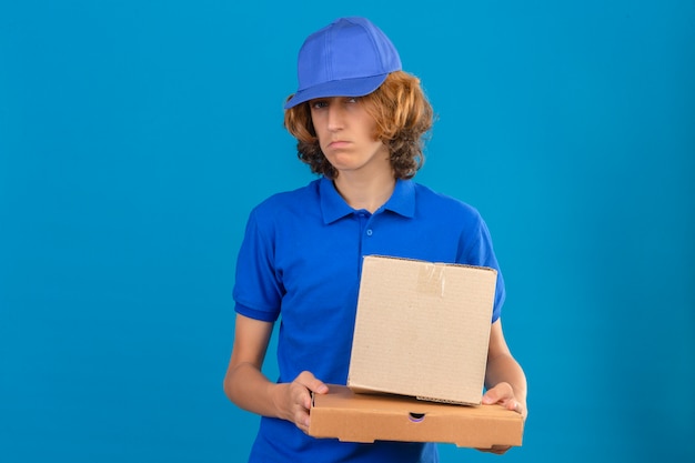 無料写真 青いポロシャツと分離の青い背景の上に立って悲しそうな表情で段ボール箱を保持しているキャップを着ている若い配達人