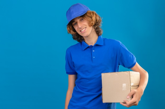 파란색 폴로 셔츠와 격리 된 파란색 배경 위에 친절한 서 웃 고 손에 골 판지 상자를 들고 모자를 입고 젊은 배달 남자