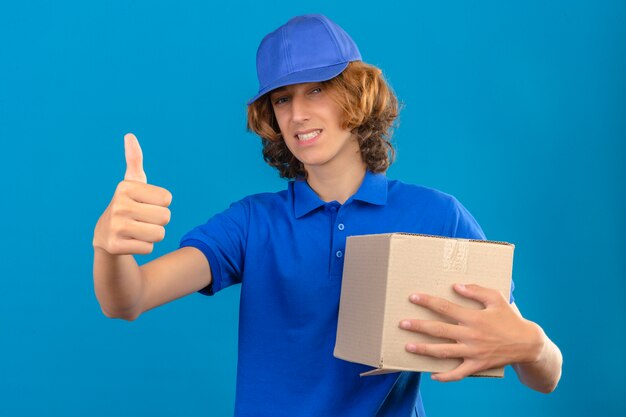 Young delivery man wearing blue polo shirt and cap holding cardboard box showing thumb up smiling cheerfully over isolated blue background