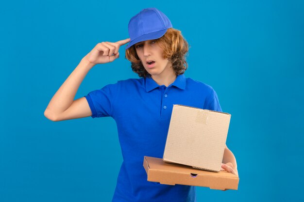 Young delivery man wearing blue polo shirt and cap holding cardboard boxes pointing to head with one finger great idea or thought standing over isolated blue background