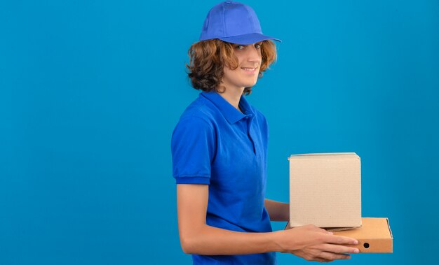 青いポロシャツと分離の青い背景の上にずるずる立っている笑顔段ボール箱を保持しているキャップを着ている若い配達人
