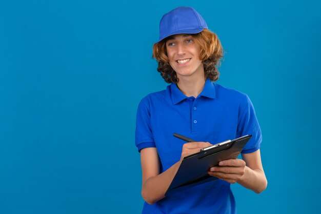 Молодой курьер в синей рубашке поло и кепке держит в руках буфер обмена, что-то пишет, весело улыбаясь на синем фоне