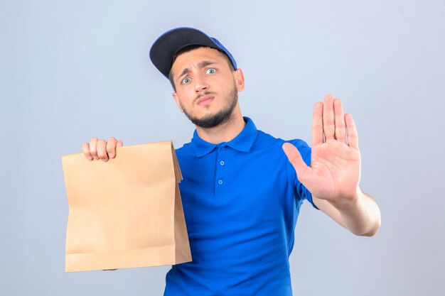 Young delivery man wearing blue polo shirt and cap holding paper bag with takeaway food looking worried making stop gesture with hand over isolated white background