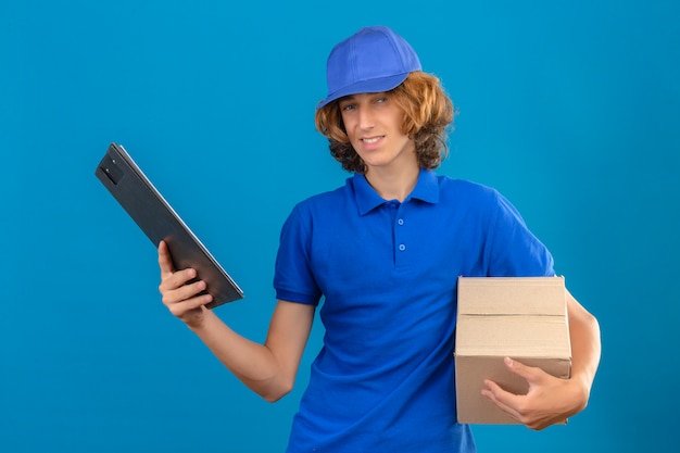 Young delivery man wearing blue polo shirt and cap holding pizza box and clipboard with blanks while smiling friendly standing with happy face over isolated blue background