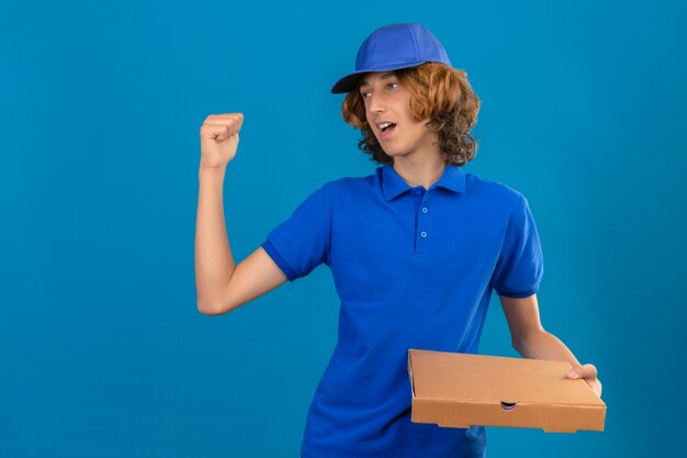 青いポロシャツとピザのボックスを保持している若い配達人が手と親指で分離された青い背景に不満を探して親指で後ろを後ろに指しているピザの箱を保持