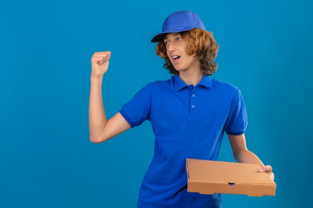 Young delivery man wearing blue polo shirt and cap holding pizza box pointing to the back behind with hand and thumb up looking dissatisfied over isolated blue background