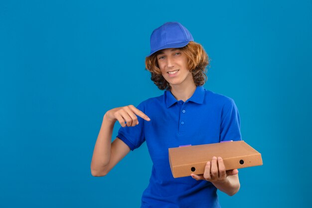Young delivery man wearing blue polo shirt and cap holding pizza box pointing index finger to box while looking at camera with big smile on face standing over isolated blue background