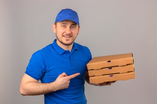 Young delivery man wearing blue polo shirt and cap holding stack of pizza boxes pointing finger at them looking at camera with smile standing over isolated white background