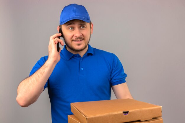 Young delivery man wearing blue polo shirt and cap holding stack of pizza boxes while talking on mobile phone looking nervous and confused over isolated white background