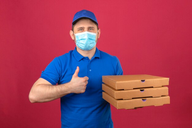 Молодой курьер в синей рубашке поло и кепке в медицинской маске, держа стопку коробок для пиццы, показывая большой палец вверх со счастливым лицом на изолированном розовом фоне