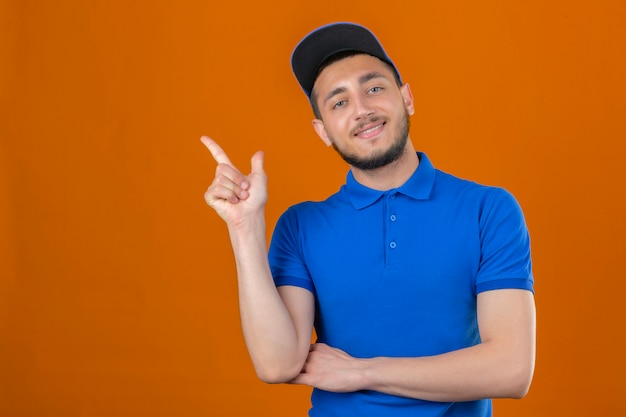 파란색 폴로 셔츠와 격리 된 오렌지 배경 위에 자신감과 행복을 찾고 측면에 검지 손가락으로 가리키는 모자를 착용하는 젊은 배달 남자
