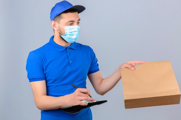 파란색 폴로 셔츠와 보호 의료 마스크를 착용하는 젊은 배달 남자는 고객에게 패키지를 제공하고 격리 된 흰색 배경 위에 클립 보드에 쓰기