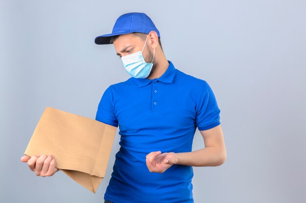 Молодой курьер в синей рубашке поло и кепке в защитной медицинской маске, стоящий с бумажным пакетом, сомневающийся с запутанным выражением лица на изолированном белом фоне