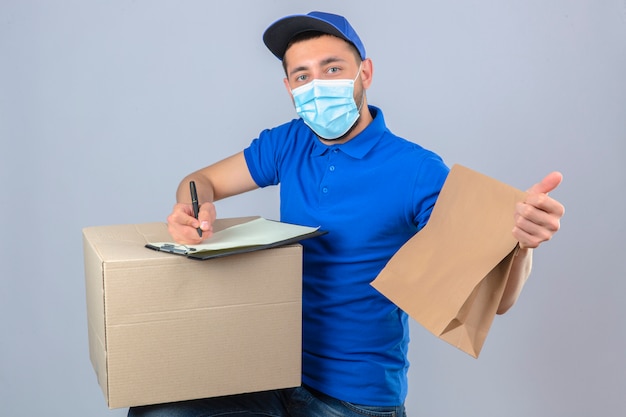 孤立した白い背景の上のクリップボードのボックスに紙のパッケージの書き込みと防護マスク立って青いポロシャツとキャップを着ている若い配達人