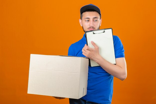 Young delivery man wearing blue polo shirt and cap standing with big cardboard box and clipboard looking away over isolated orange background