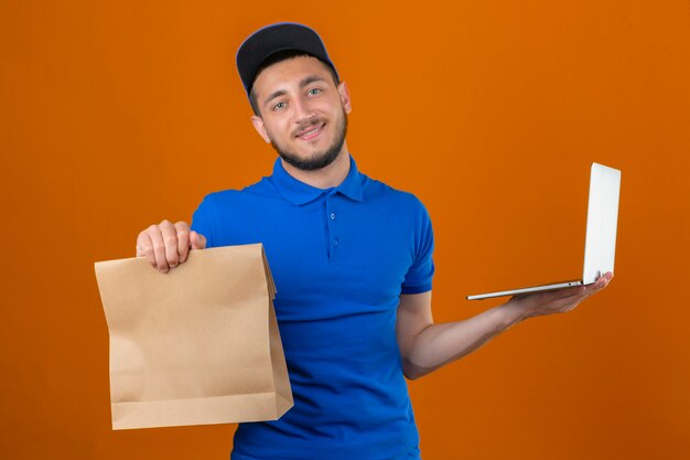 Young delivery man wearing blue polo shirt and cap standing with clipboard and paper package looking at camera with smile on face over isolated orange background