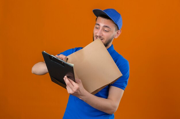 Young delivery man wearing blue polo shirt and cap standing with clipboard and paper package writing with smile on face over isolated orange background