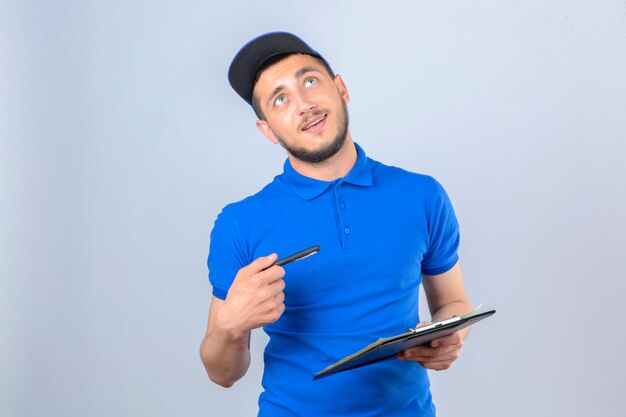 青いポロシャツとクリップボードと分離の白い背景の上の顔に笑顔で見上げるペンで立っているキャップを着ている若い配達人