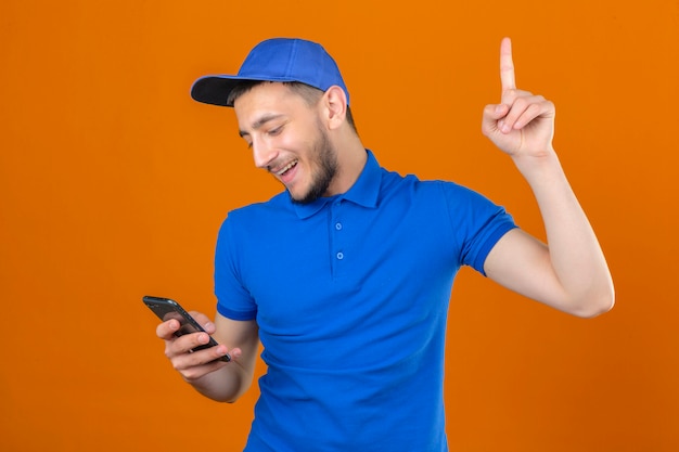 青いポロシャツと分離されたオレンジ色の背景に新しいアイデアの概念を笑顔で人差し指を手にスマートフォンで立っているキャップを着ている若い配達人