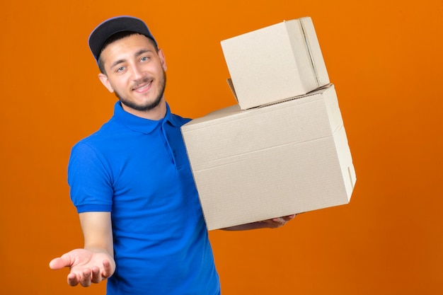 Young delivery man wearing blue polo shirt and cap standing with stack of boxes looking at camera with smile waiting payment for delivery over isolated orange background