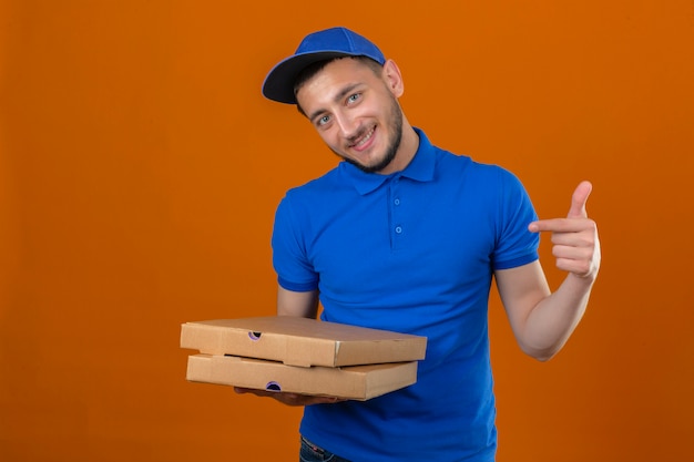 Young delivery man wearing blue polo shirt and cap standing with stack of pizza boxes looking at camera with smile on face pointing with finger over isolated orange background