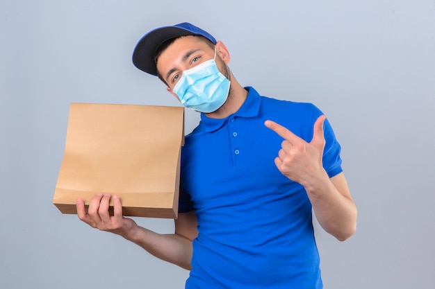 Young delivery man wearing blue polo shirt and cap wearing protective medical mask holding paper package with takeaway food pointing to this package with finger smiling over isolated white backgrou