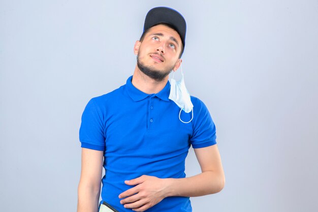 Young delivery man wearing blue polo shirt and cap with medical protective mask on ear staring up thinking with smile over isolated white background