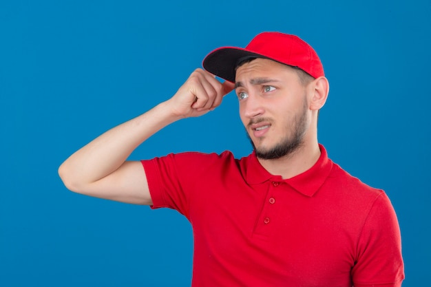 young delivery man wearing red polo shirt and cap confused feels doubtful and unsure over isolated blue background
