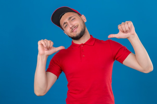 Young delivery man wearing red polo shirt and cap looking at camera smiling cheerfully proud and self-satisfied over isolated blue background